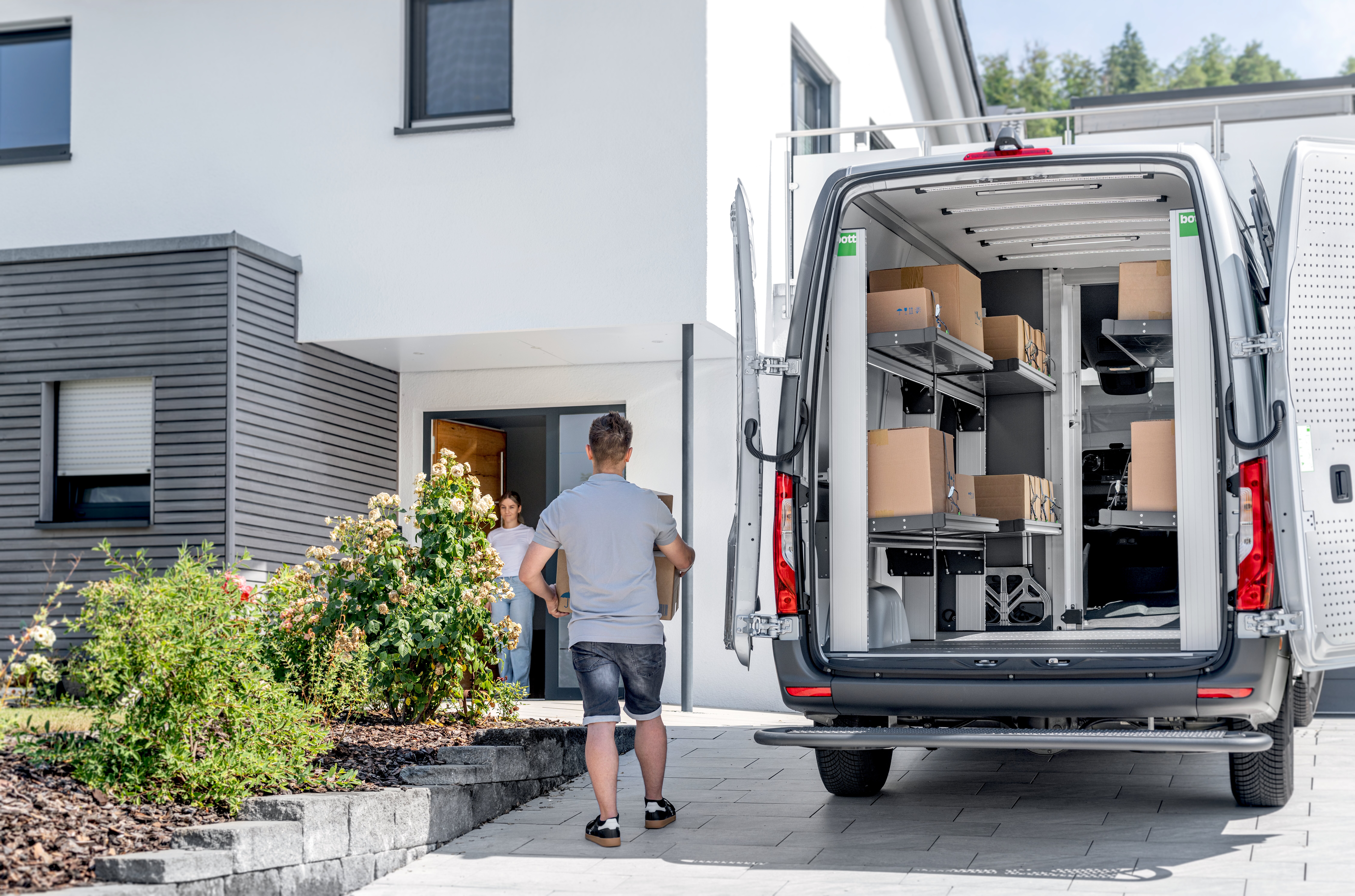 KEP-Zusteller bringt ein Paket vom Lieferfahrzeug zu einem Wohnhaus; im Fahrzeug sind bott vario3 Klappregale sichtbar